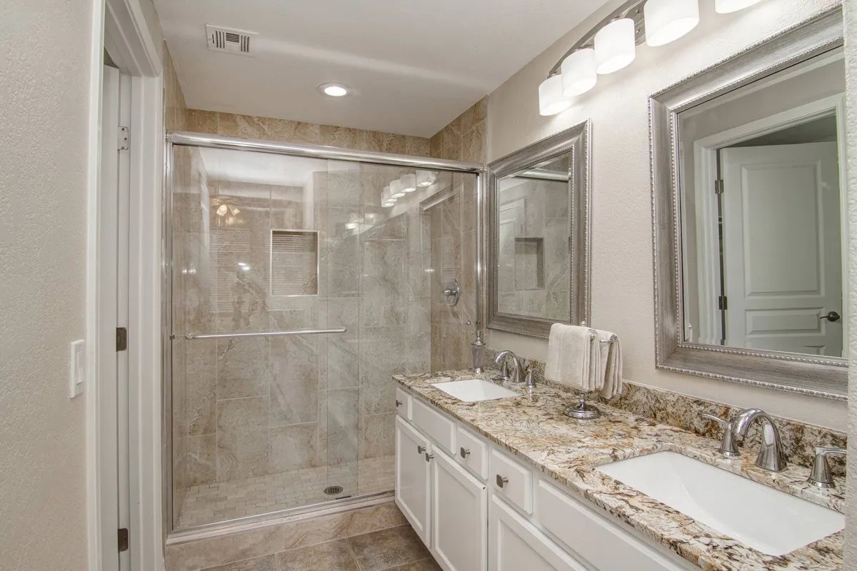 A bright, modern master bath with a glass-enclosed walk-in shower, granite vanity countertop with double sinks, stylish mirrors, and contemporary lighting fixtures. The design emphasizes both luxury and functionality, perfect for morning routines or evening relaxation.