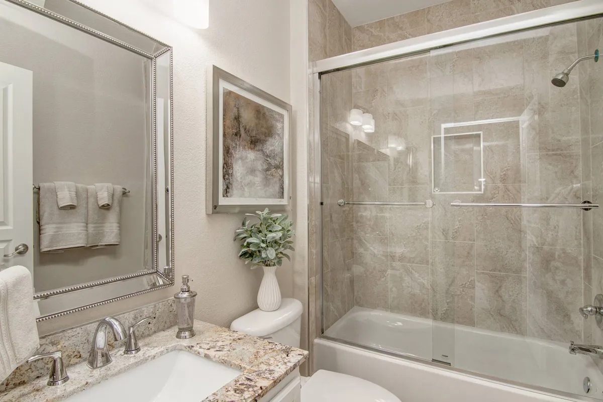 A well-lit secondary bathroom featuring a granite countertop, large mirror, shower area with a tub/shower combo, and decorative accents like an art piece and a potted plant. The space is inviting and functional, offering guests convenience and privacy.