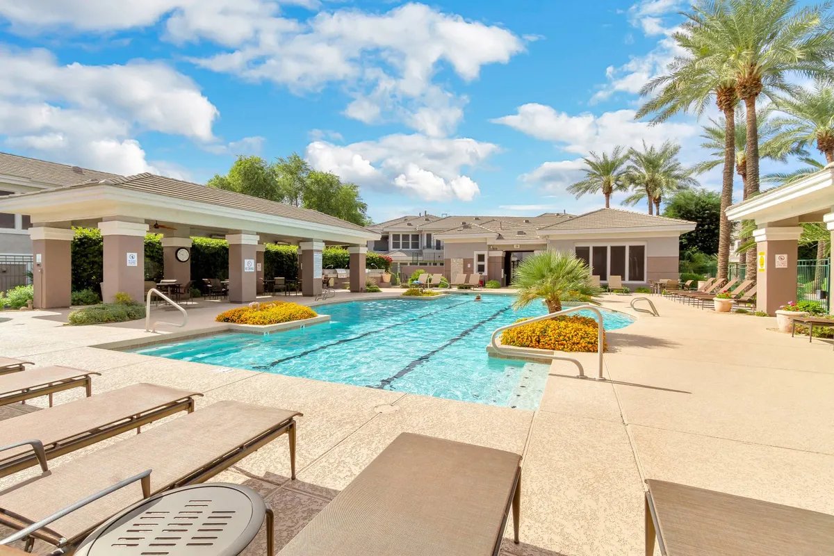 A vibrant exterior scene featuring a sparkling swimming pool surrounded by lounge chairs, lush landscaped gardens, and tall palm trees. The area is designed for leisure with ample space for sunbathing, swimming, and social gatherings.