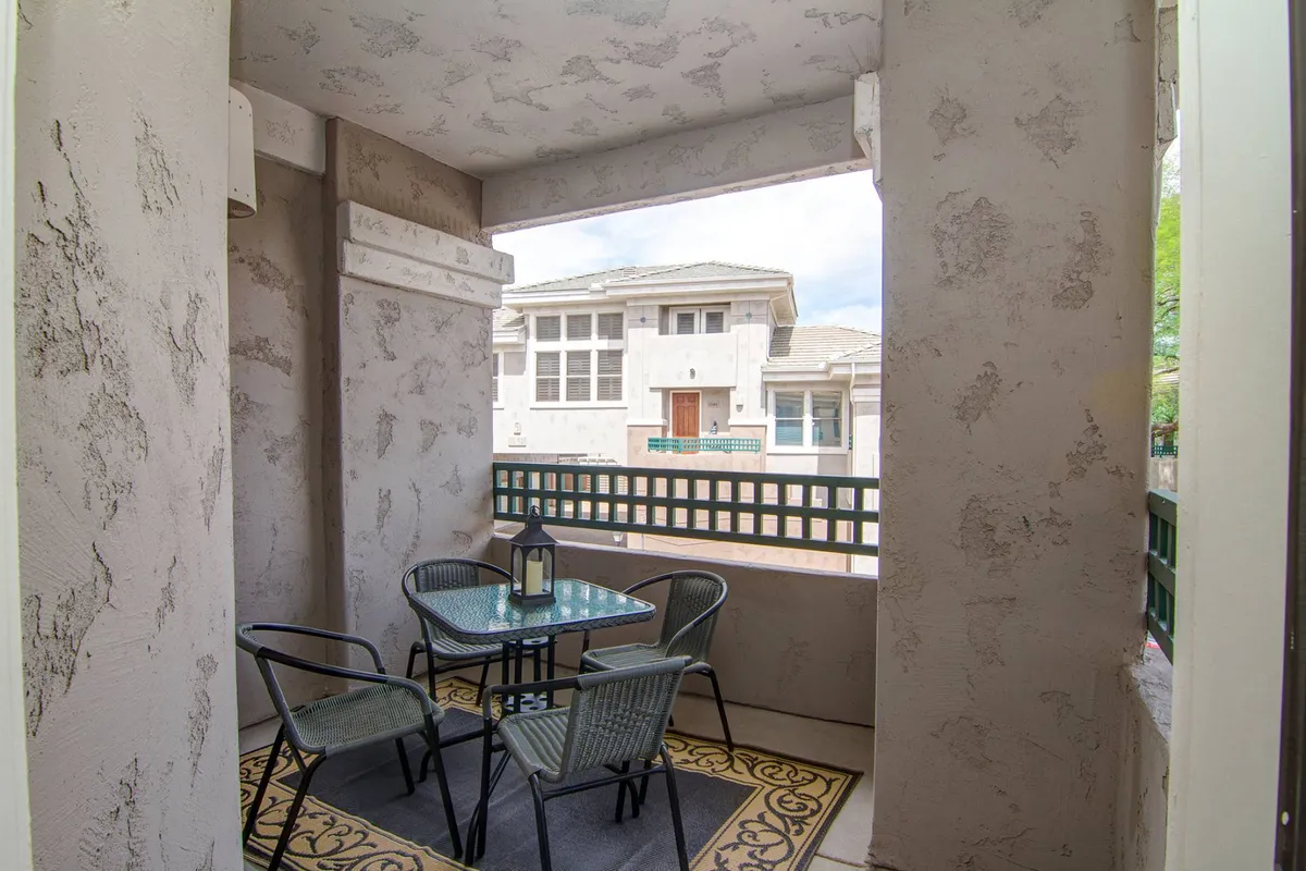 A calm, cozy balcony scene with a glass table, four chairs, a lantern centerpiece, and a decorative rug. The outdoor setting is designed for relaxed morning coffee or evening drinks, with the potential for sunset views.