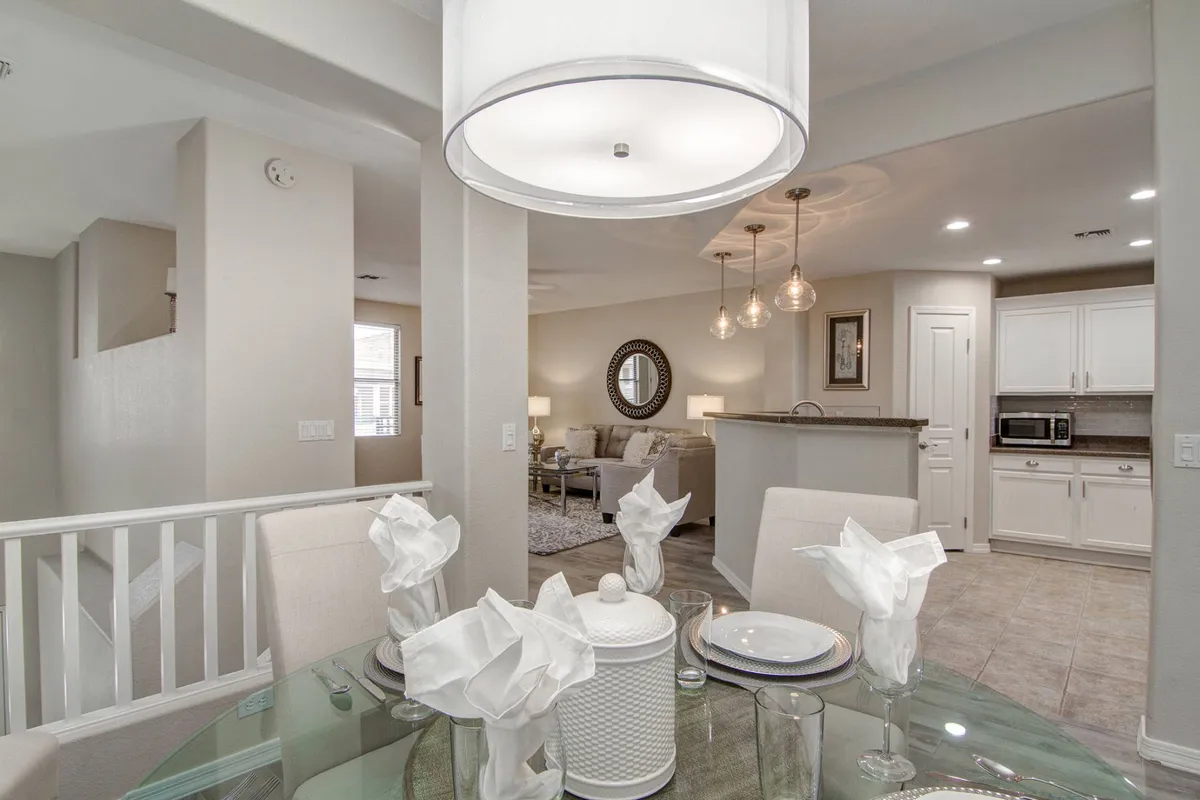 This image captures an open living area featuring a glass dining table set for four, a modern kitchen with high-end appliances and barstool seating, and a living space with a cozy couch and accent lighting. Large windows allow natural light to flood the space, while the open layout visually connects the kitchen, dining, and living areas.