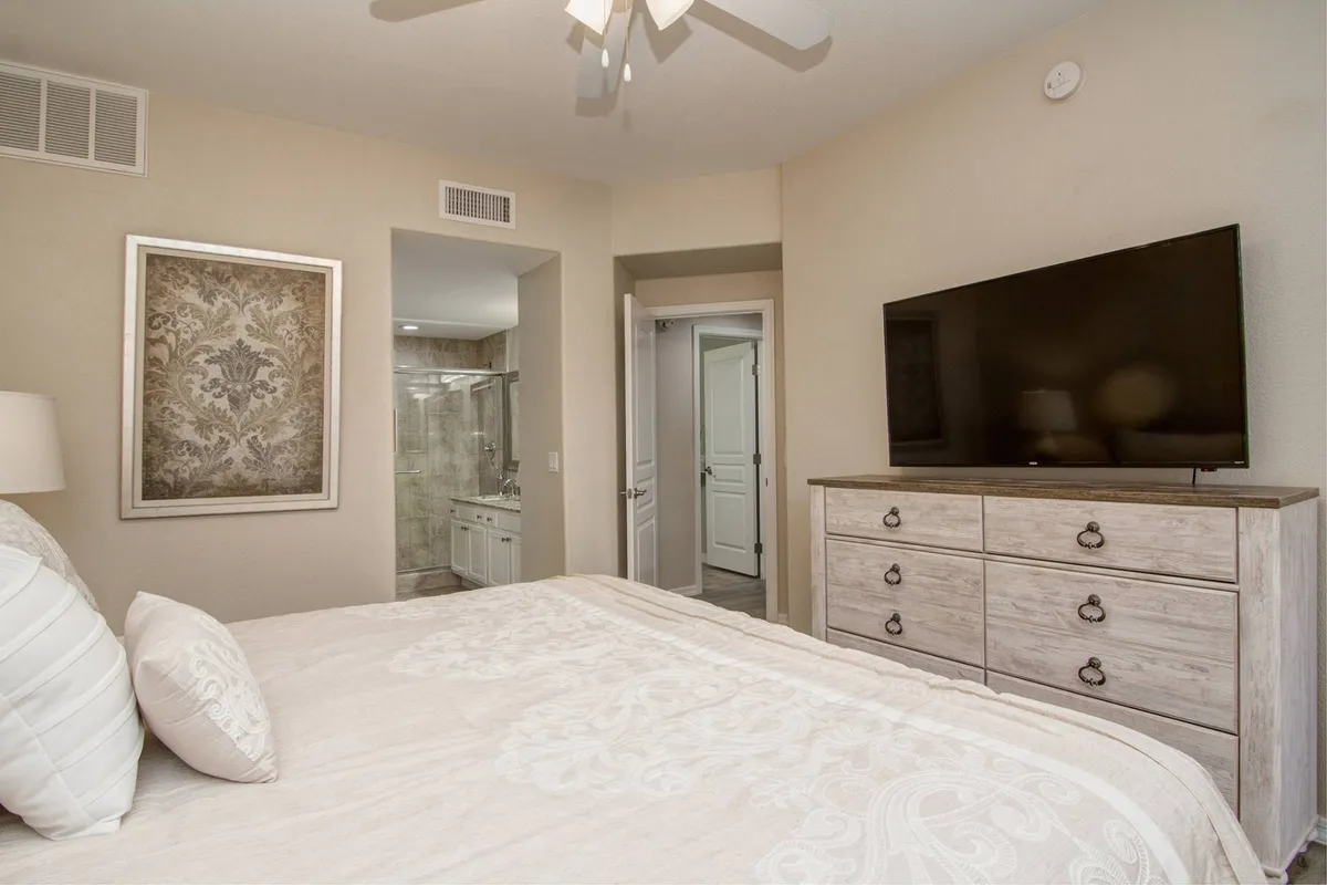 A serene and comfortable guest bedroom with a plush bed dressed in decorative pillows and soft linens, a large wall-mounted TV, framed artwork, and a well-appointed dresser. The room offers a restful atmosphere, ideal for guests seeking privacy and comfort.