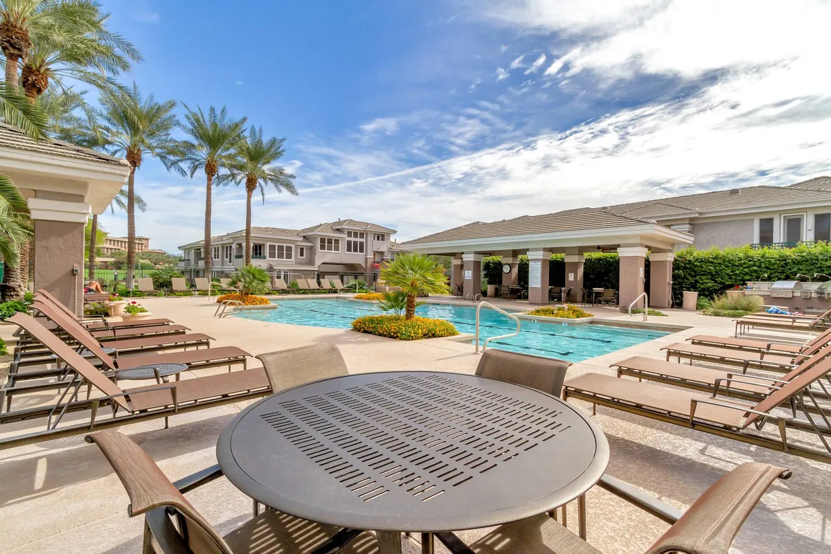 An inviting exterior scene featuring a sparkling swimming pool with lounge chairs and a nearby outdoor dining table, framed by tall palm trees. The ambiance is one of comfort and sociability, ideal for sunbathing or enjoying a meal after a round of golf.
