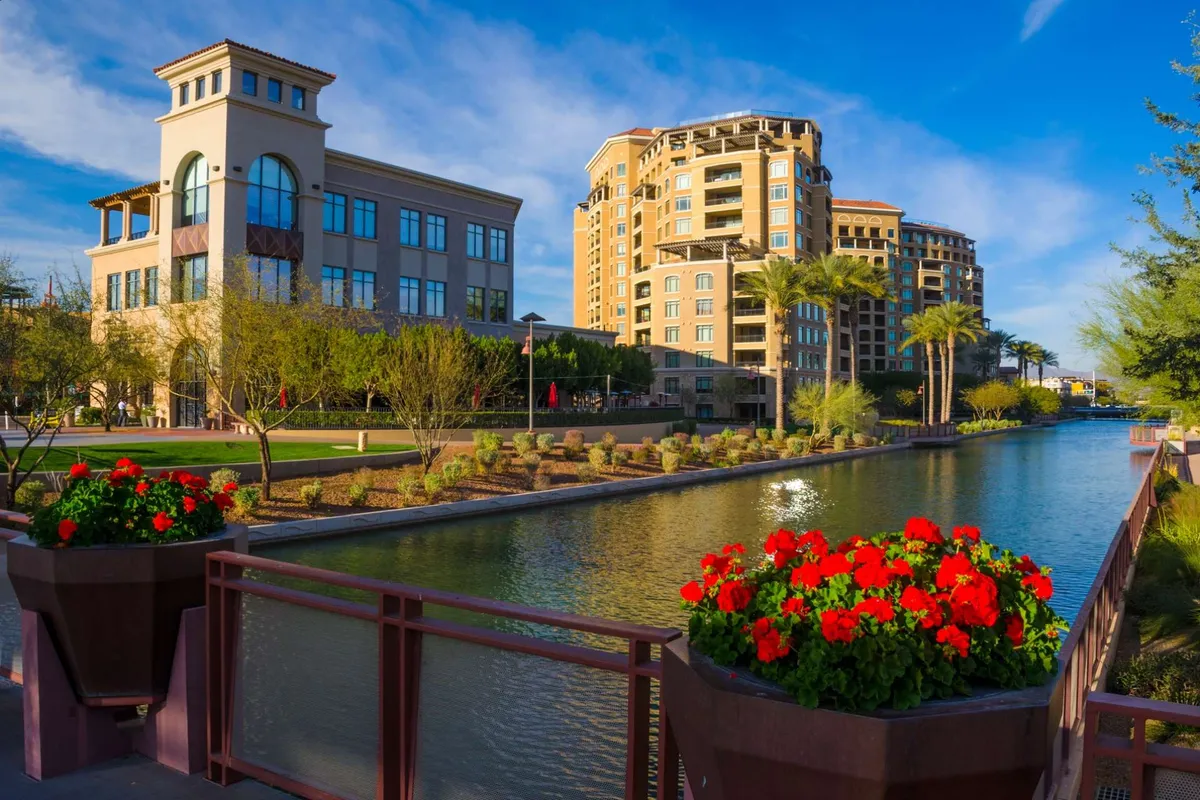 This vibrant exterior image captures a canal lined with colorful floral planters, palm trees, and nearby buildings. People can stroll or relax by the water, soaking in the lively ambiance of the area.