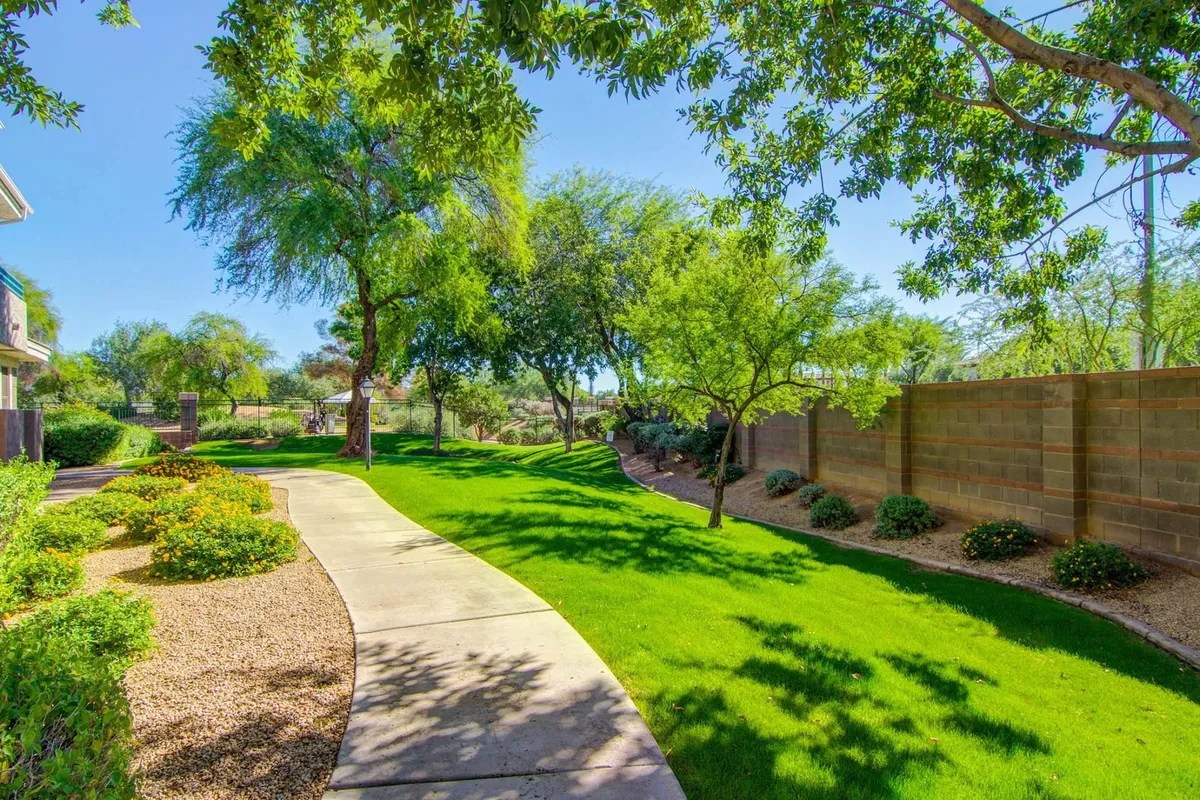 A serene exterior view of a lush green lawn with a winding pathway, shaded by mature trees and bordered by a low wall. The setting is calm and inviting, perfect for a walk or quiet outdoor relaxation between golf outings.